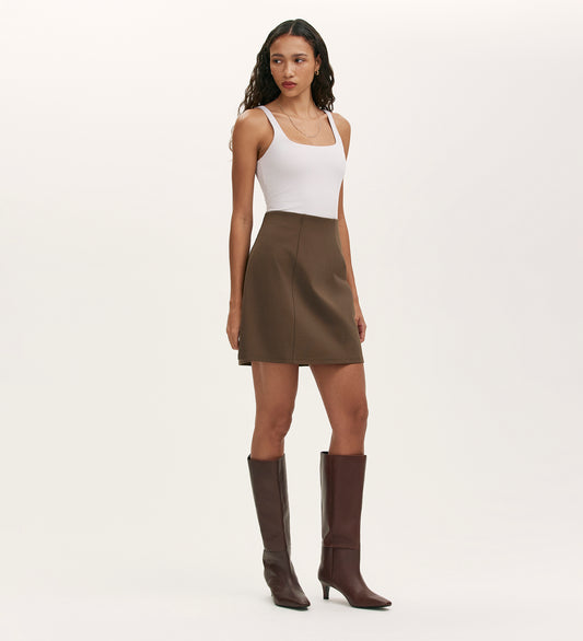 A woman with long wavy hair stands against a plain light background, wearing a white sleeveless top, knee-high brown boots, and the Helia Mole Ponte Jersey Midi Skirt with an elasticated high waist, facing slightly to the side.