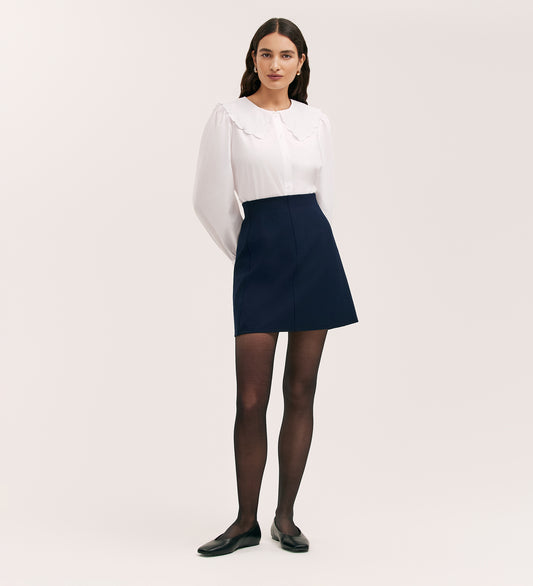A woman faces forward wearing a white long-sleeve blouse with a scalloped collar, the Helia Navy Ponte Jersey Midi Skirt featuring an elasticated waistband, black tights, and black flats against a plain light background.