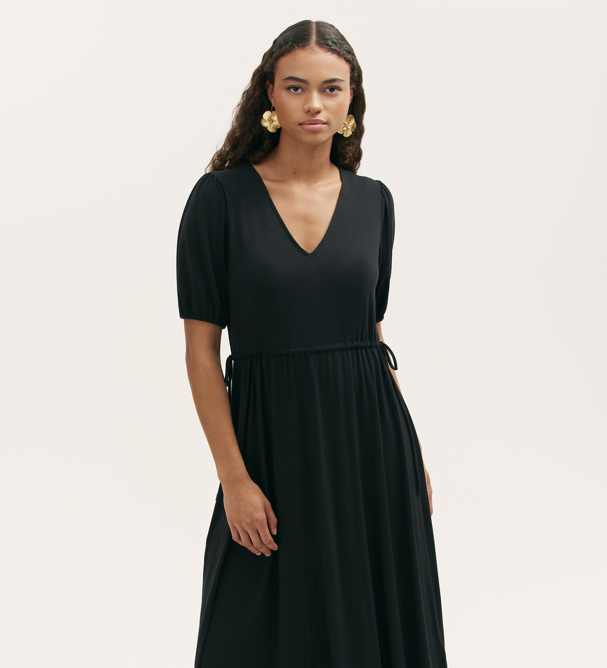 A woman with long curly hair wears gold earrings and the Hessa Black Jersey Midi Dress with a flattering fit-and-flare silhouette, standing against a plain light background.