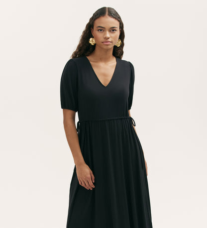 A woman with long curly hair wears gold earrings and the Hessa Black Jersey Midi Dress with a flattering fit-and-flare silhouette, standing against a plain light background.