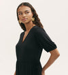 A woman with long curly hair models the Hessa Black Jersey Midi Dress—featuring a flattering V-neck, short sleeves, and fit-and-flare silhouette—styled with large gold flower earrings against a plain light background.