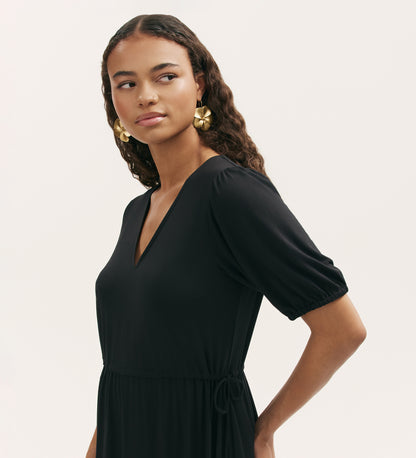 A woman with long curly hair models the Hessa Black Jersey Midi Dress—featuring a flattering V-neck, short sleeves, and fit-and-flare silhouette—styled with large gold flower earrings against a plain light background.