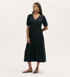 A woman in the Hessa Black Jersey Midi Dress, featuring a flattering fit-and-flare silhouette and V-neckline, stands against a plain light background. She pairs it with gold earrings and tan sandals for a versatile look.