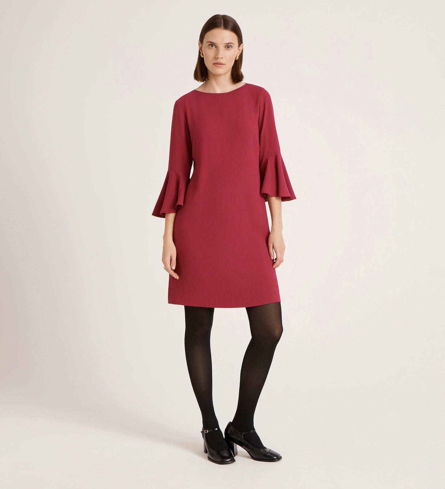 A woman stands against a plain background, wearing the Izzy Berry Crepe Short Dress with bell sleeves, black tights, and shoes. She has straight, shoulder-length hair and a neutral expression.
