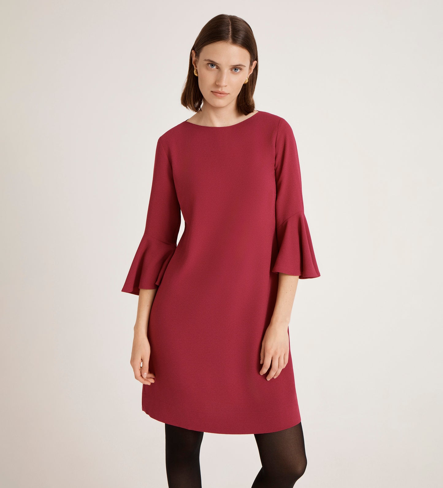 A woman with straight brown hair wears the Izzy Berry Crepe Short Dress, knee-length in dark pink with ruffled cuffs. She stands facing forward against a plain light background, her expression neutral.