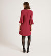 A woman with shoulder-length brown hair faces away, wearing a knee-length Izzy Berry Crepe Short Dress with bell sleeves in dark pink, paired with black tights and heeled shoes against a plain white background.
