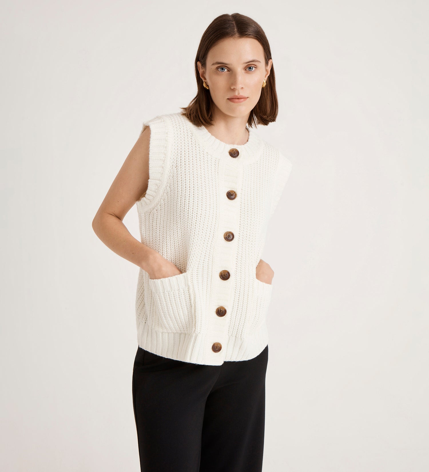 A woman with straight, shoulder-length brown hair models the Janette Ivory Knitted Vest—a sleeveless white vest with large brown buttons and pockets—paired with black pants, set against a plain light background.