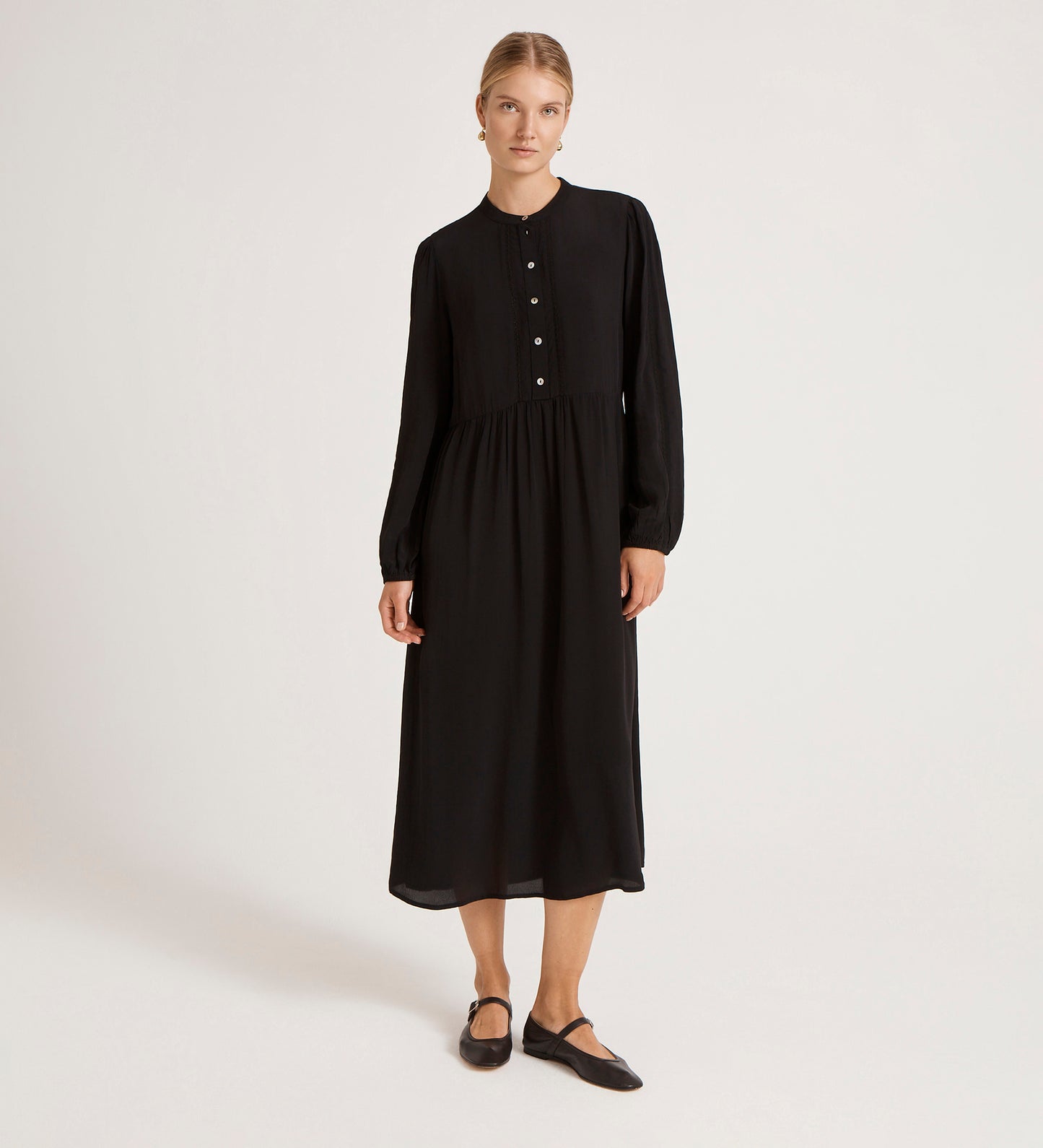 A woman stands against a plain background wearing the Jasmine Black Lace Trim Crepe Midi Dress with long sleeves and button details. She pairs it with black flats, her hair pulled back, and her arms relaxed by her sides.