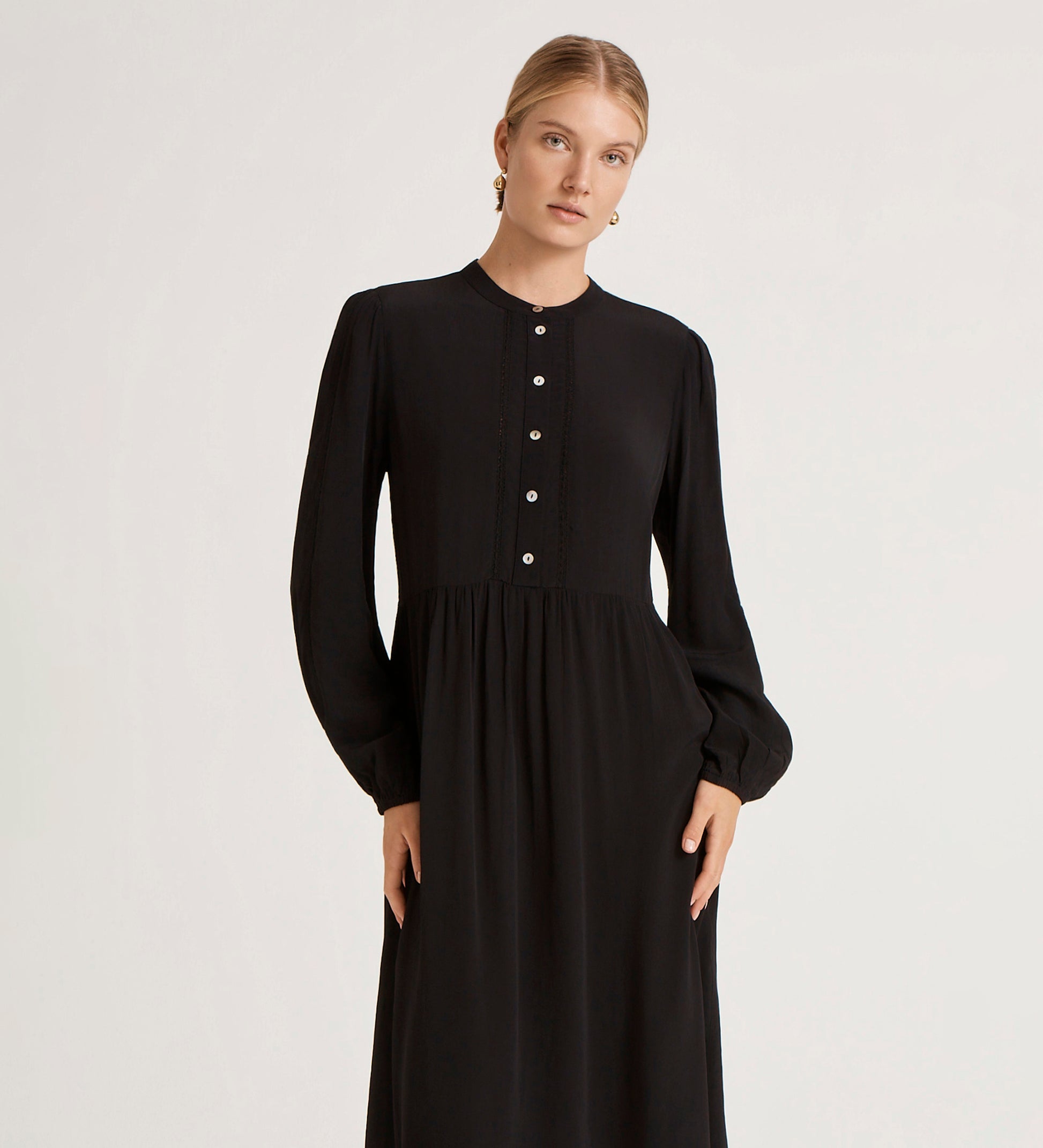 A woman stands calmly against a plain light background, wearing the Jasmine Black Lace Trim Crepe Midi Dress with a vintage-inspired silhouette and looking at the camera.