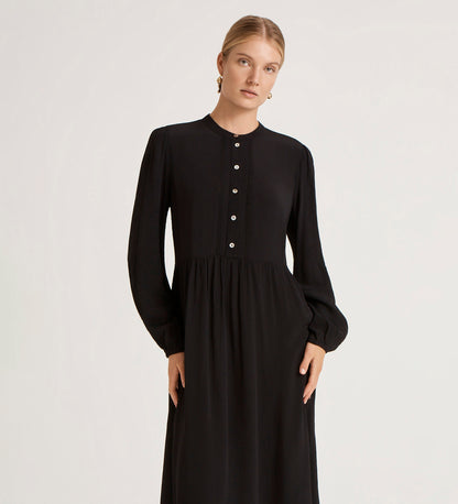 A woman stands calmly against a plain light background, wearing the Jasmine Black Lace Trim Crepe Midi Dress with a vintage-inspired silhouette and looking at the camera.