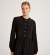 A woman with blonde hair tied back is wearing the Jasmine Black Lace Trim Crepe Midi Dress, featuring long sleeves, a mandarin collar, buttoned front, and gold earrings, standing against a plain light background.