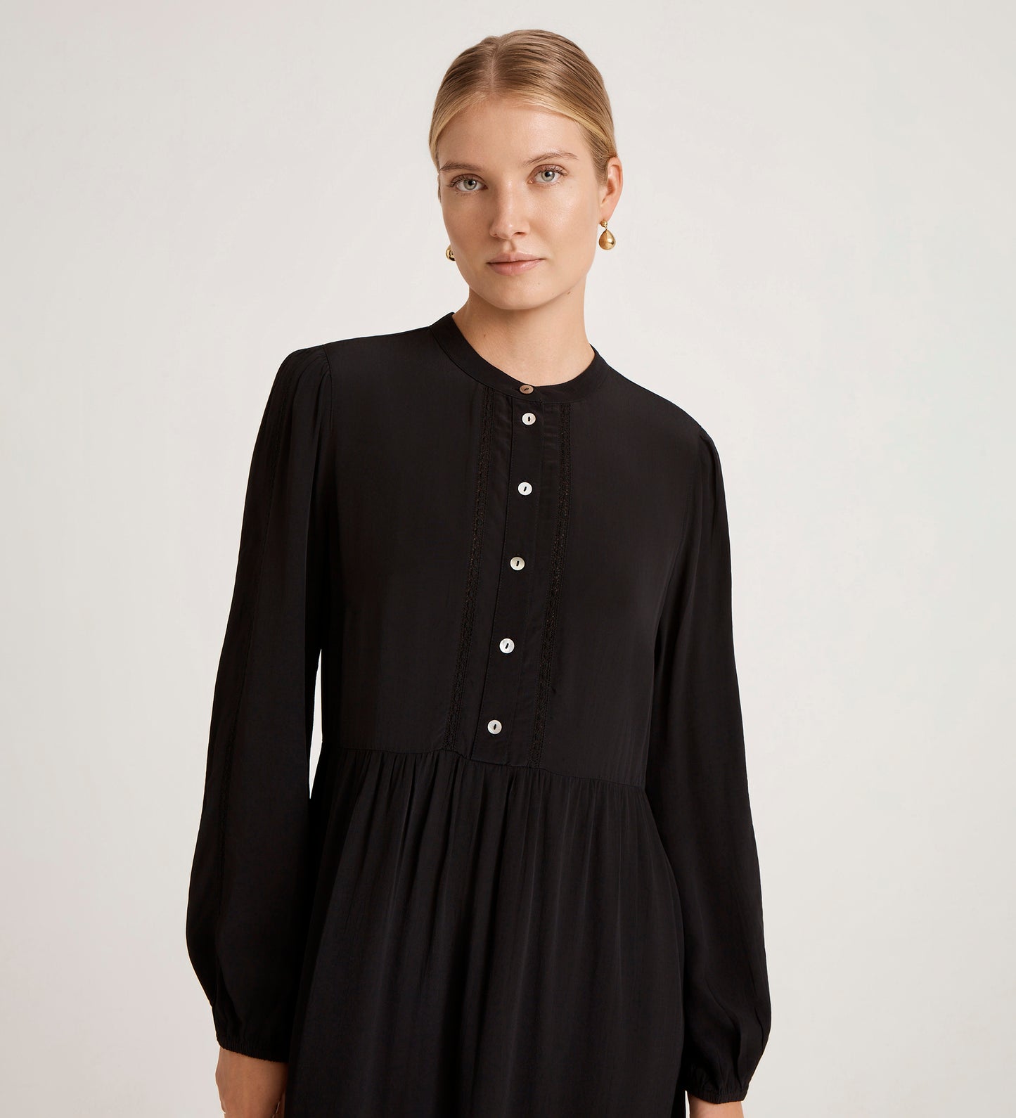 A woman with blonde hair tied back is wearing the Jasmine Black Lace Trim Crepe Midi Dress, featuring long sleeves, a mandarin collar, buttoned front, and gold earrings, standing against a plain light background.