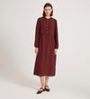 A woman stands against a plain background wearing the Jasmine Burgundy Lace Trim Crepe Midi Dress, featuring long sleeves, a gathered waist, and lace trim. She pairs it with brown flats and has straight brown hair and a neutral expression.