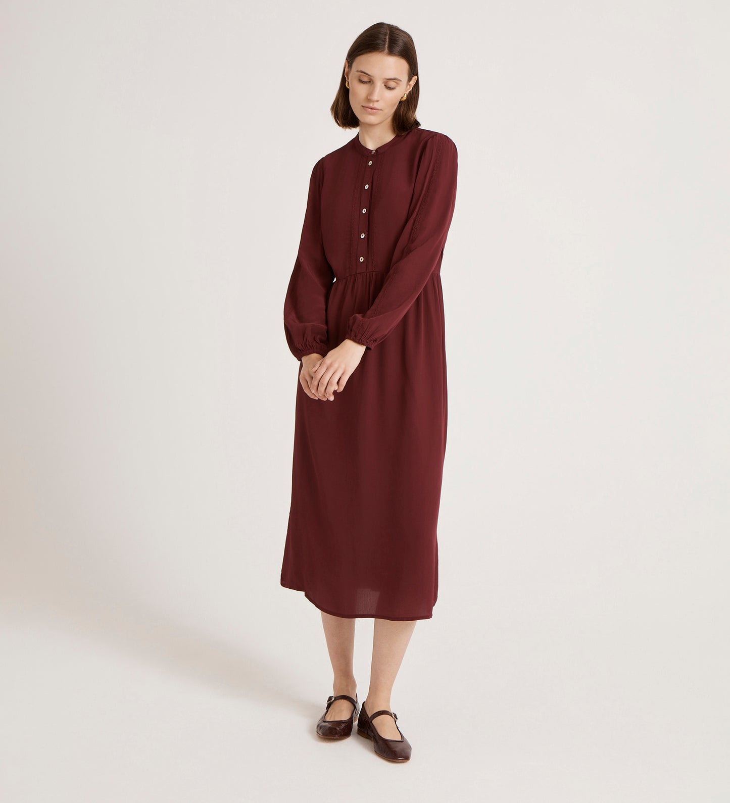 A woman stands against a plain background wearing the Jasmine Burgundy Lace Trim Crepe Midi Dress with long sleeves and a mandarin collar, paired with brown flat shoes. She has straight shoulder-length hair, looks down, and her hands are loosely clasped.