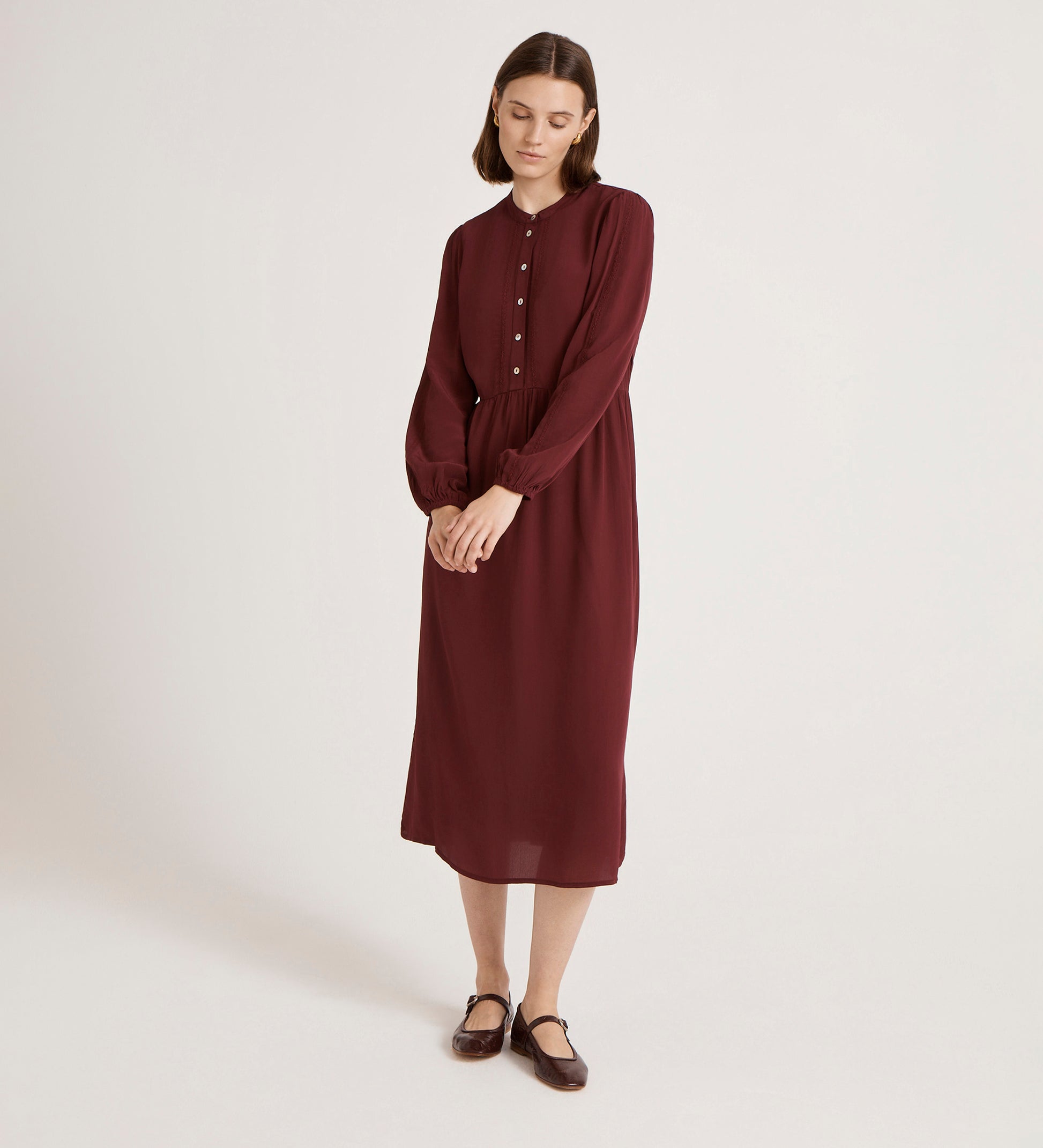 A woman stands against a plain background wearing the Jasmine Burgundy Lace Trim Crepe Midi Dress with long sleeves and a mandarin collar, paired with brown flat shoes. She has straight shoulder-length hair, looks down, and her hands are loosely clasped.