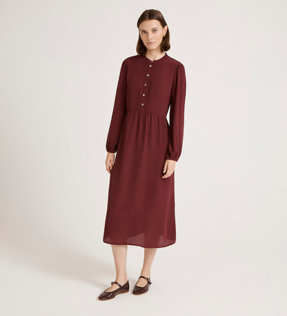 A woman with shoulder-length brown hair stands against a plain background, wearing the Jasmine Burgundy Lace Trim Crepe Midi Dress with long sleeves, button front, and gathered waist, paired with brown strap shoes.