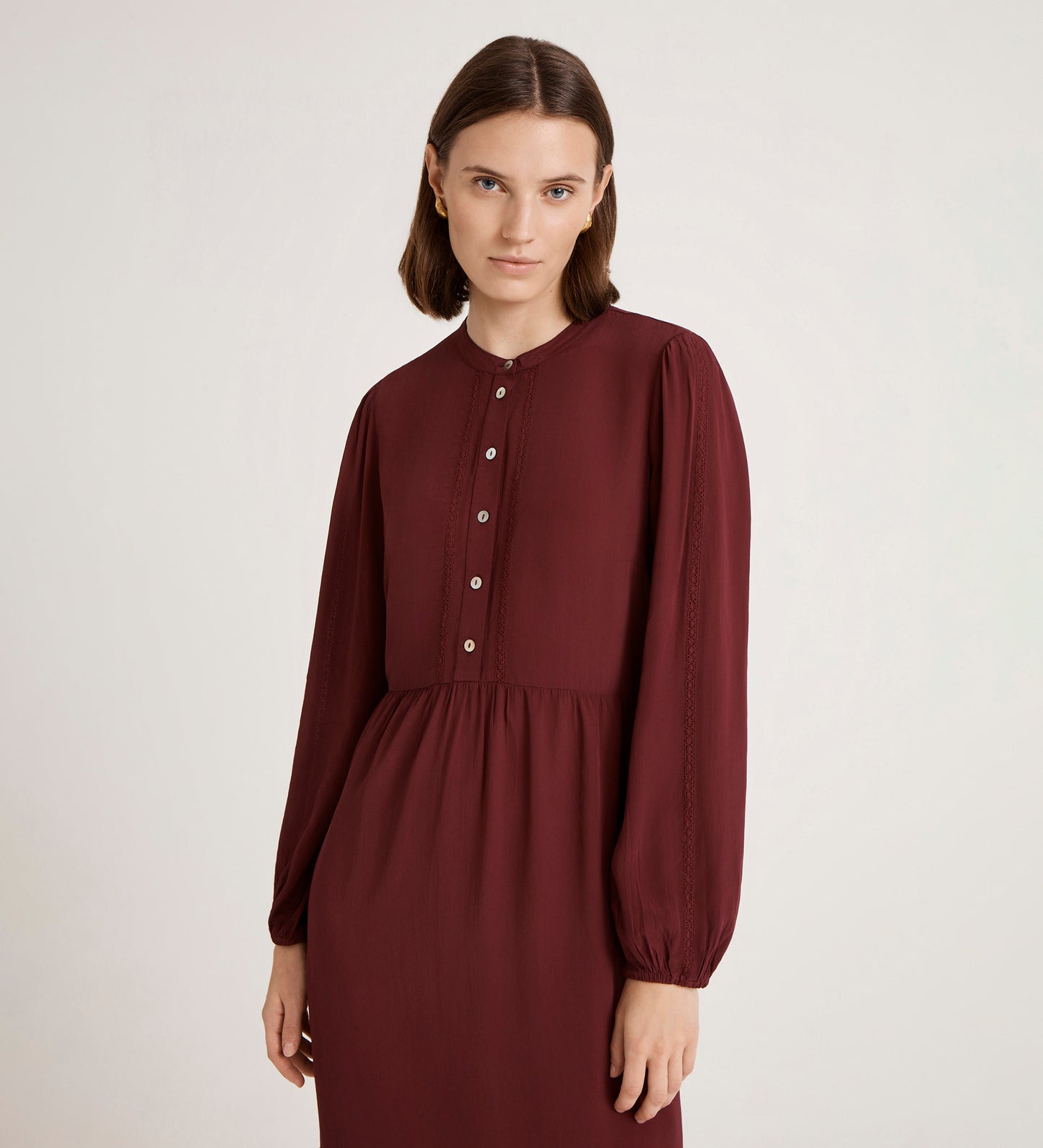 A woman with straight brown hair wears the Jasmine Burgundy Lace Trim Crepe Midi Dress, featuring front buttons, as she stands against a plain light background and gazes softly at the camera.