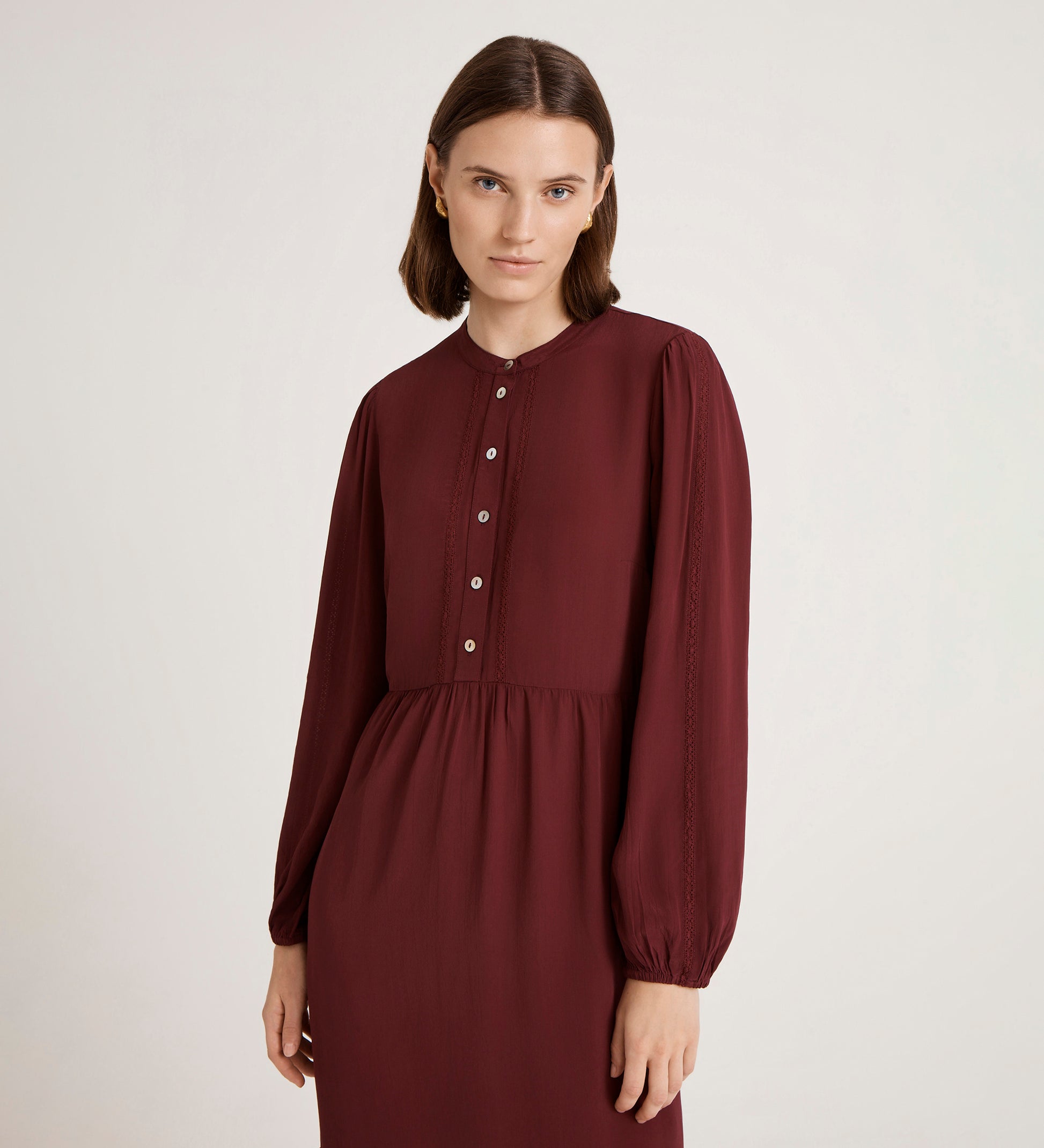 A woman with straight brown hair wears the Jasmine Burgundy Lace Trim Crepe Midi Dress, featuring front buttons, as she stands against a plain light background and gazes softly at the camera.