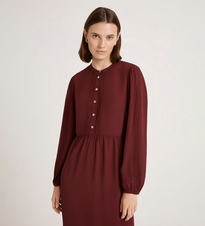 A woman with straight brown hair wears the Jasmine Burgundy Lace Trim Crepe Midi Dress, featuring front buttons, as she stands against a plain light background and gazes softly at the camera.
