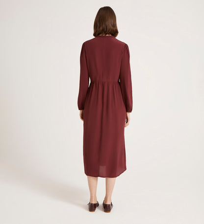 A person faces away from the camera, wearing the Jasmine Burgundy Lace Trim Crepe Midi Dress with long sleeves and brown shoes, set against a plain light background.