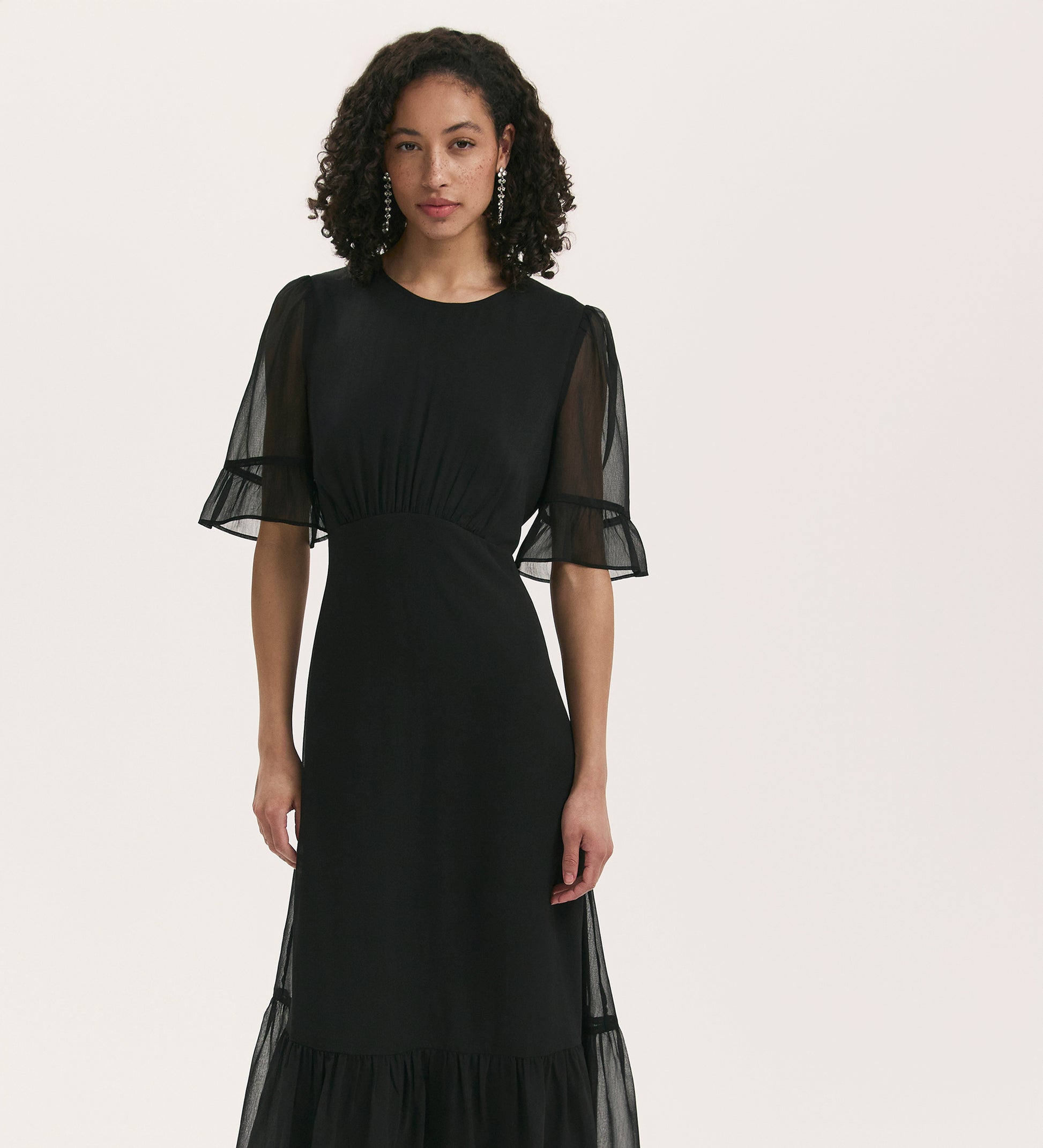 A woman with curly hair wears the Jennifer Black Chiffon Midi Dress, featuring a round neck, sheer short sleeves, and a ruffled hem, standing against a plain light background.