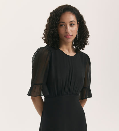 A woman with curly hair wears the Jennifer Black Chiffon Midi Dress, featuring sheer, puffed short sleeves and ruffled cuffs, paired with pearl drop earrings. She stands against a plain light background, gazing softly to the side.