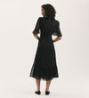 A woman with curly hair faces away, wearing the Jennifer Black Chiffon Midi Dress featuring sheer flutter sleeves and a ruffled hem, paired with black flats, set against a plain light background.