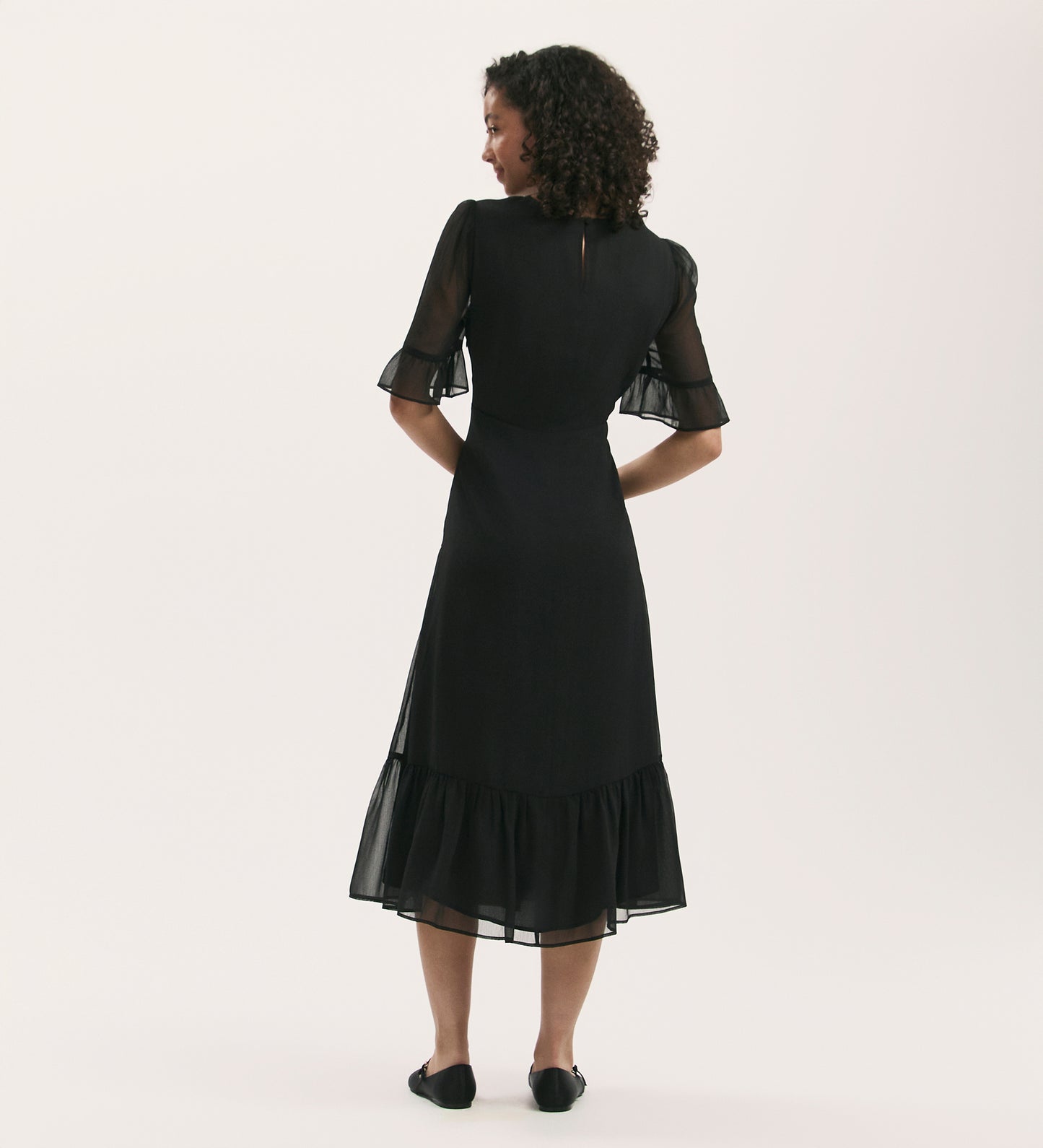A woman with curly hair faces away, wearing the Jennifer Black Chiffon Midi Dress featuring sheer flutter sleeves and a ruffled hem, paired with black flats, set against a plain light background.