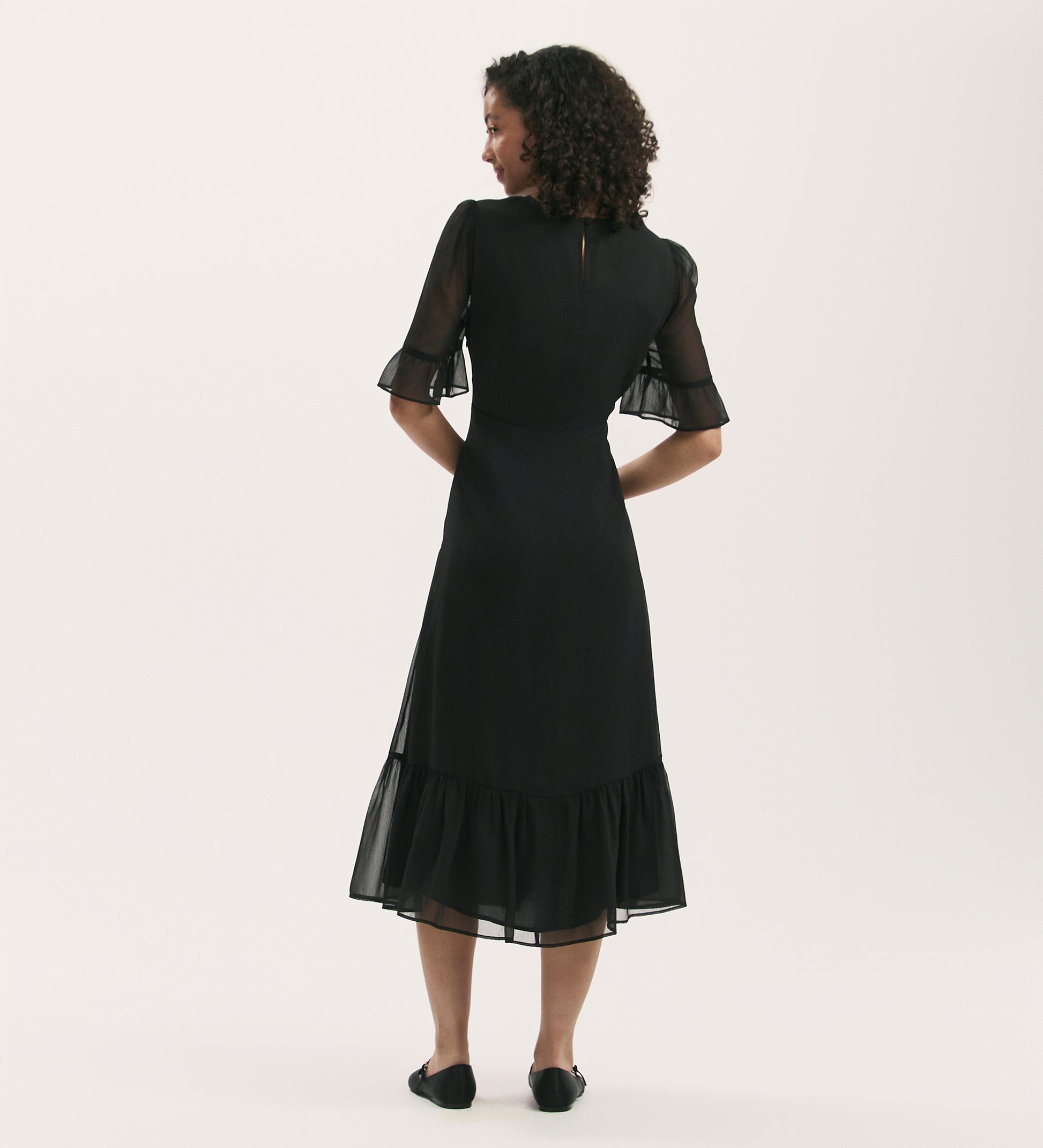 A woman with curly hair faces away, wearing the Jennifer Black Chiffon Midi Dress featuring sheer flutter sleeves and a ruffled hem, paired with black flats, set against a plain light background.