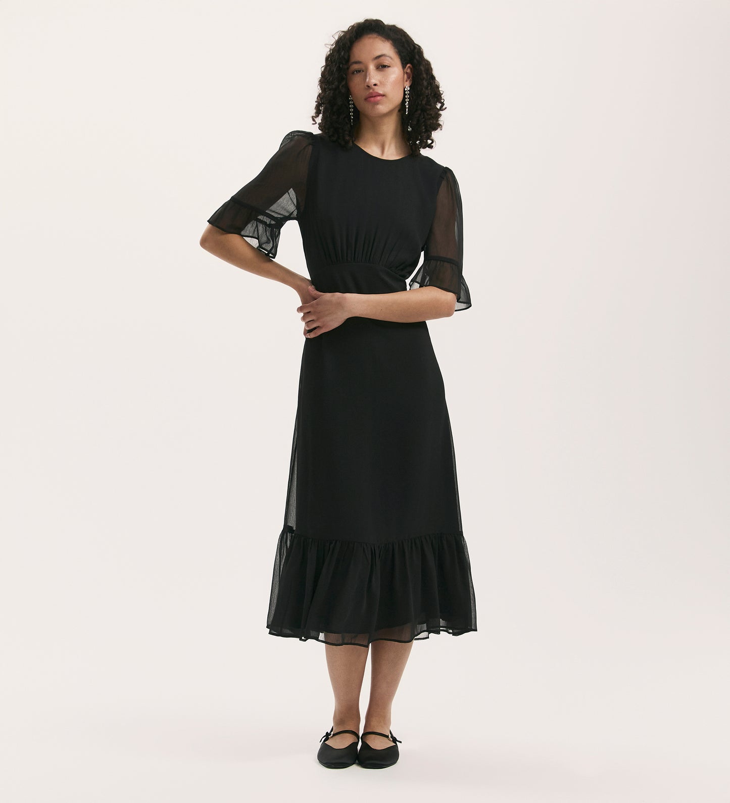 A woman with curly hair models the Jennifer Black Chiffon Midi Dress, which has sheer short sleeves, a ruffled hem, and a round neckline. She stands with arms loosely crossed against a plain white background in black flat shoes.