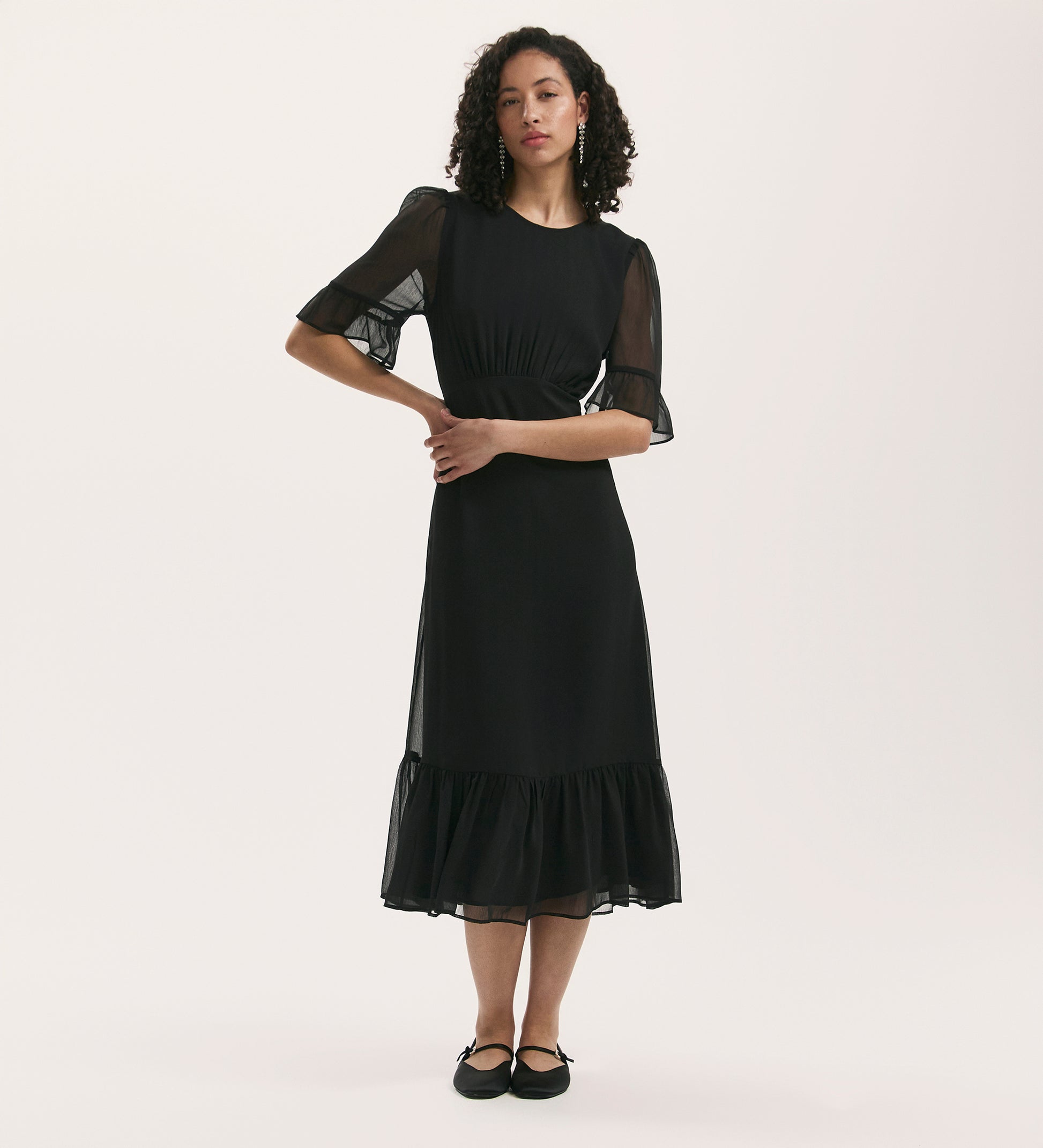 A woman with curly hair models the Jennifer Black Chiffon Midi Dress, which has sheer short sleeves, a ruffled hem, and a round neckline. She stands with arms loosely crossed against a plain white background in black flat shoes.
