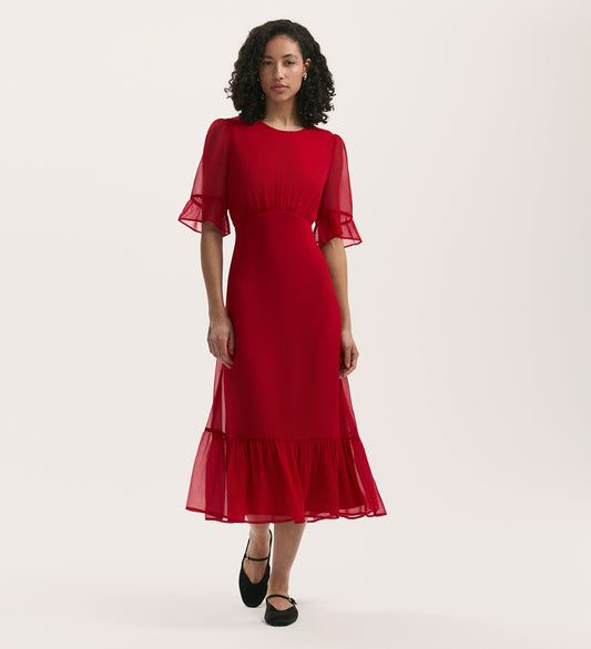 A woman with curly hair wears the Jennifer Red Chiffon Midi Dress, featuring a round neck, sheer short sleeves, and ruffled hem. She stands against a plain white background and pairs the fit and flare dress with black shoes.