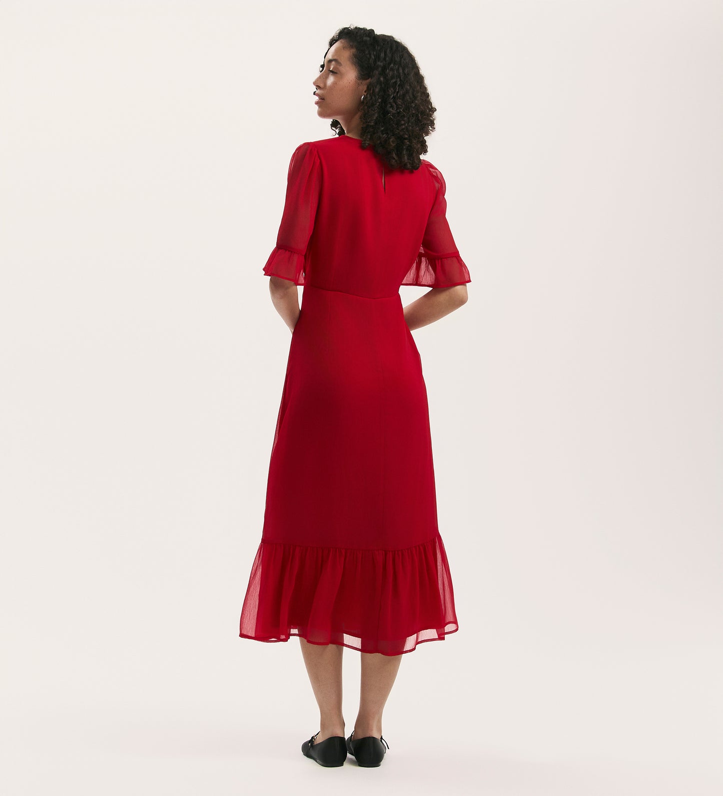A woman with curly hair faces away, wearing the Jennifer Red Chiffon Midi Dress featuring short sleeves and a ruffled hem, paired with black flat shoes, set against a plain white background.