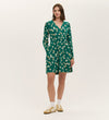 A woman stands against a plain white background wearing the Joanne Green Poppies Jersey Short Dress, which features long sleeves, a V-neckline, and a white and yellow floral pattern, paired with white socks and beige sneakers.