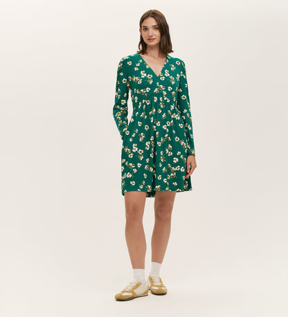 A woman stands against a plain white background wearing the Joanne Green Poppies Jersey Short Dress, which features long sleeves, a V-neckline, and a white and yellow floral pattern, paired with white socks and beige sneakers.