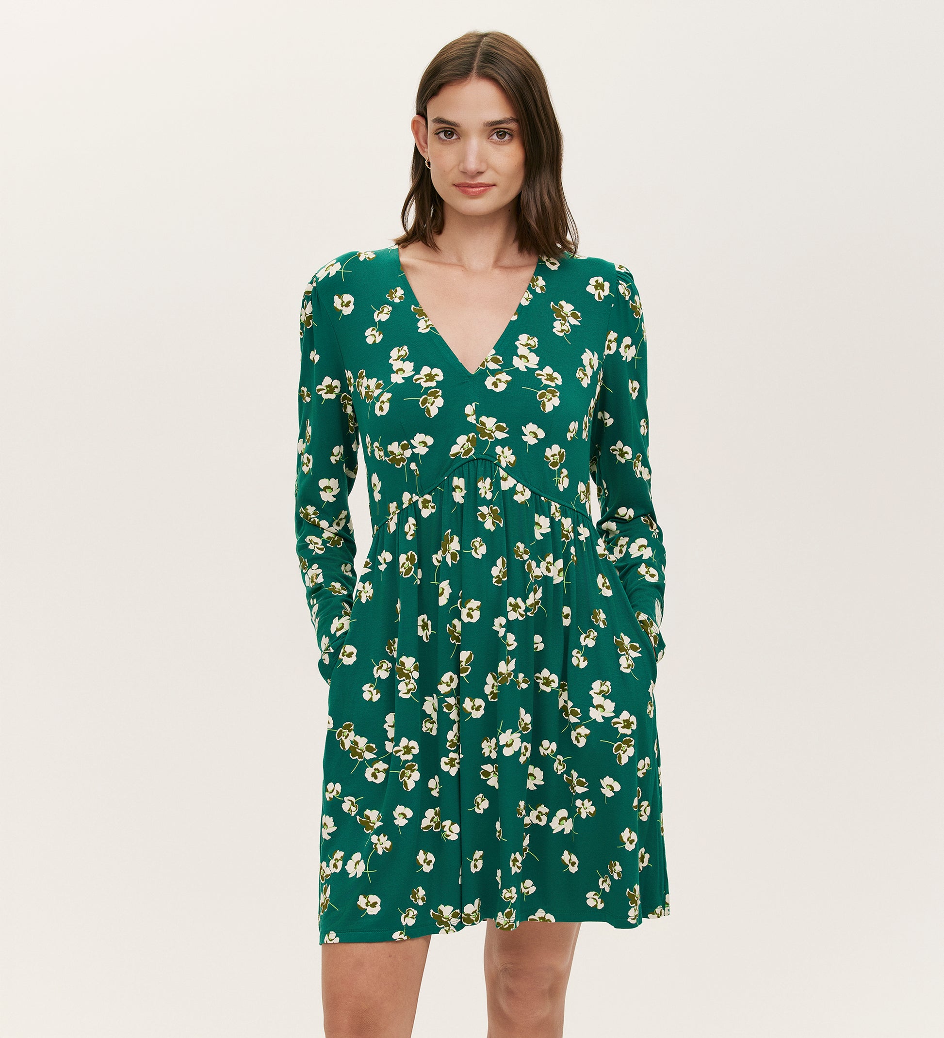 A woman in the Joanne Green Poppies Jersey Short Dress featuring long sleeves, a V-neckline, yellow and white floral print, and pockets poses against a plain light background.
