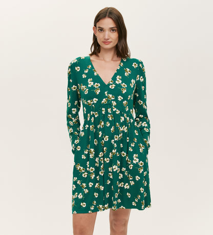 A woman in the Joanne Green Poppies Jersey Short Dress featuring long sleeves, a V-neckline, yellow and white floral print, and pockets poses against a plain light background.