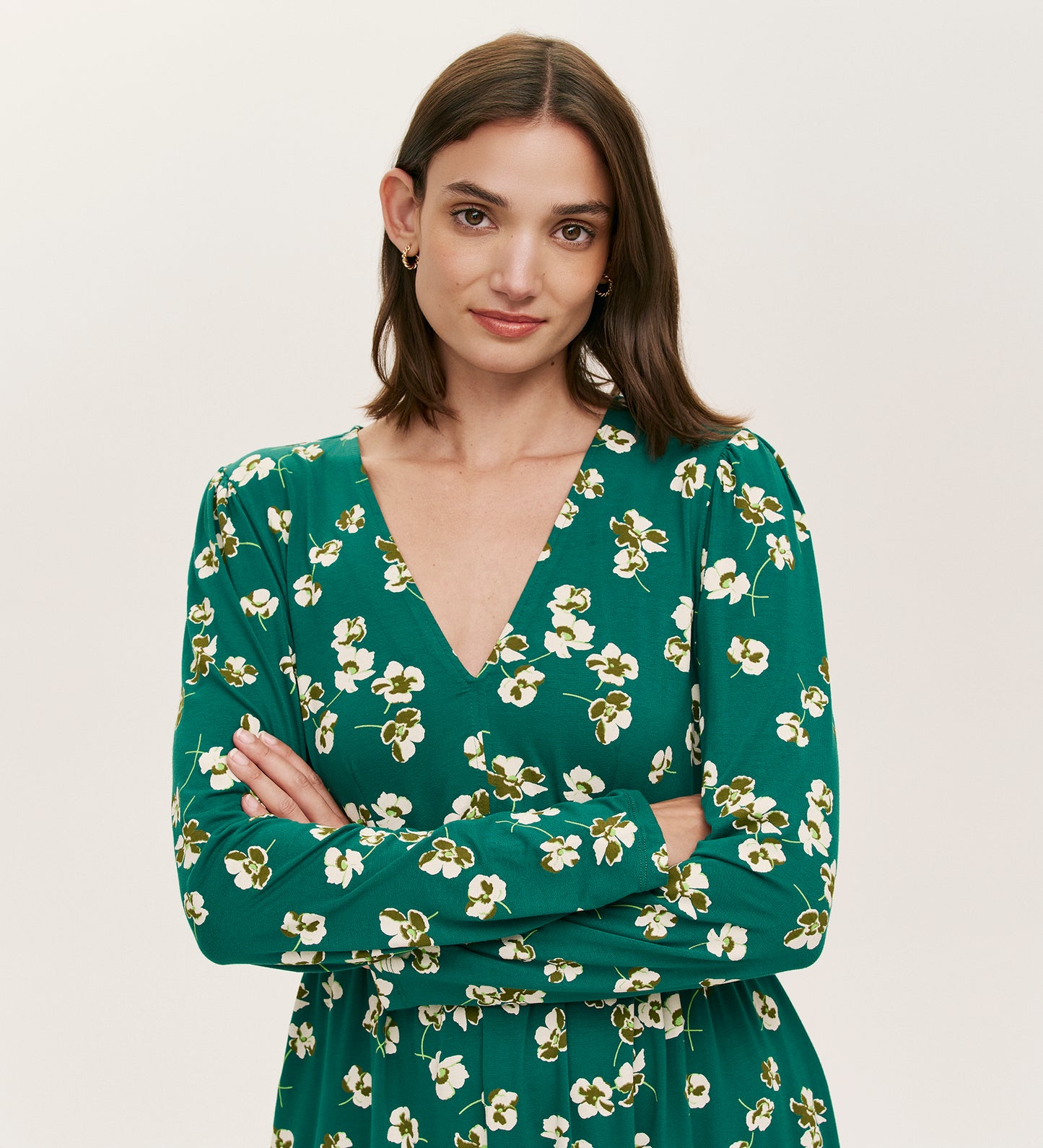 A woman with straight brown hair wears the Joanne Green Poppies Jersey Short Dress, featuring a white floral pattern and V-neckline. She stands with arms crossed, smiling slightly against a plain, light background.