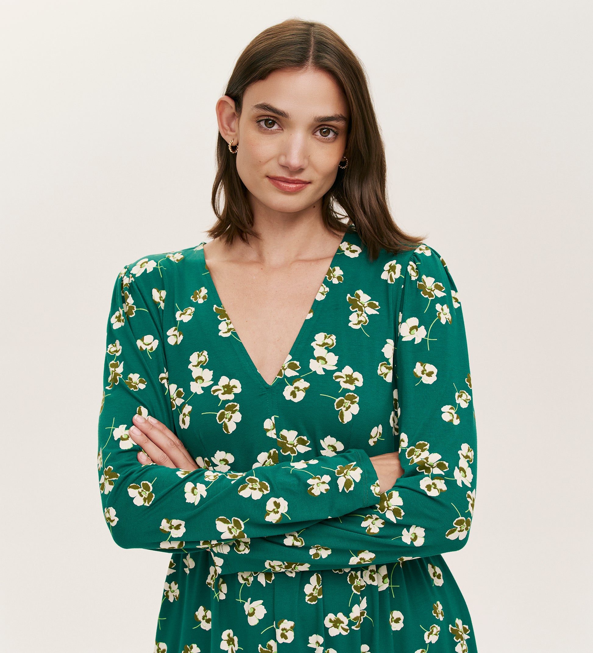 A woman with straight brown hair wears the Joanne Green Poppies Jersey Short Dress, featuring a white floral pattern and V-neckline. She stands with arms crossed, smiling slightly against a plain, light background.