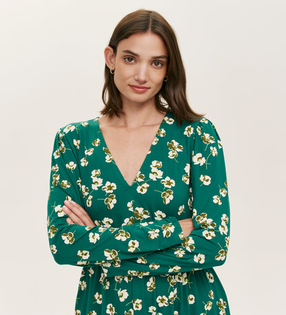 A woman with straight brown hair wears the Joanne Green Poppies Jersey Short Dress, featuring a white floral pattern and V-neckline. She stands with arms crossed, smiling slightly against a plain, light background.