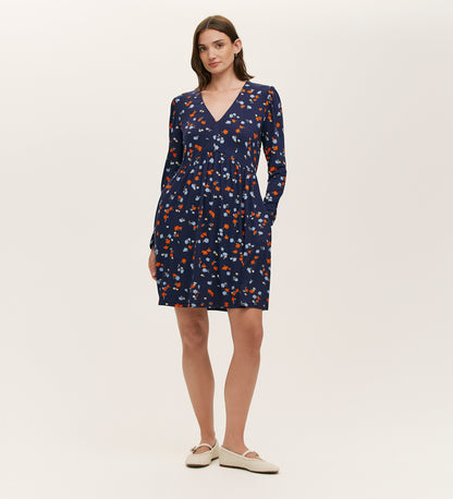 A woman wears the Joanne Navy Ditsy Jersey Short Dress, featuring long sleeves, a V-neckline, and a red, white, and orange floral print. She has straight brown hair, beige slip-on shoes, and stands against a plain light background.