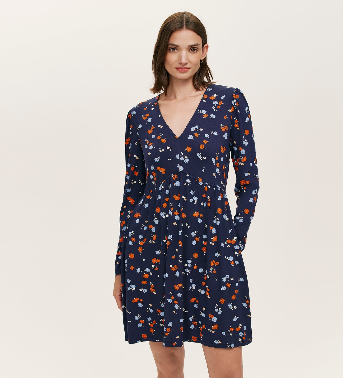 A woman models the Joanne Navy Ditsy Jersey Short Dress, featuring long sleeves, a V-neckline, and a pattern of small red, white, and light blue flowers against a plain light background.