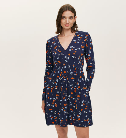 A woman models the Joanne Navy Ditsy Jersey Short Dress, featuring long sleeves, a V-neckline, and a pattern of small red, white, and light blue flowers against a plain light background.