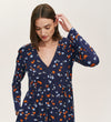 A woman with straight brown hair models the Joanne Navy Ditsy Jersey Short Dress, a mini dress with a V-neckline, long sleeves, and an orange, blue, and white floral print. She looks downward and touches her hair with one hand.
