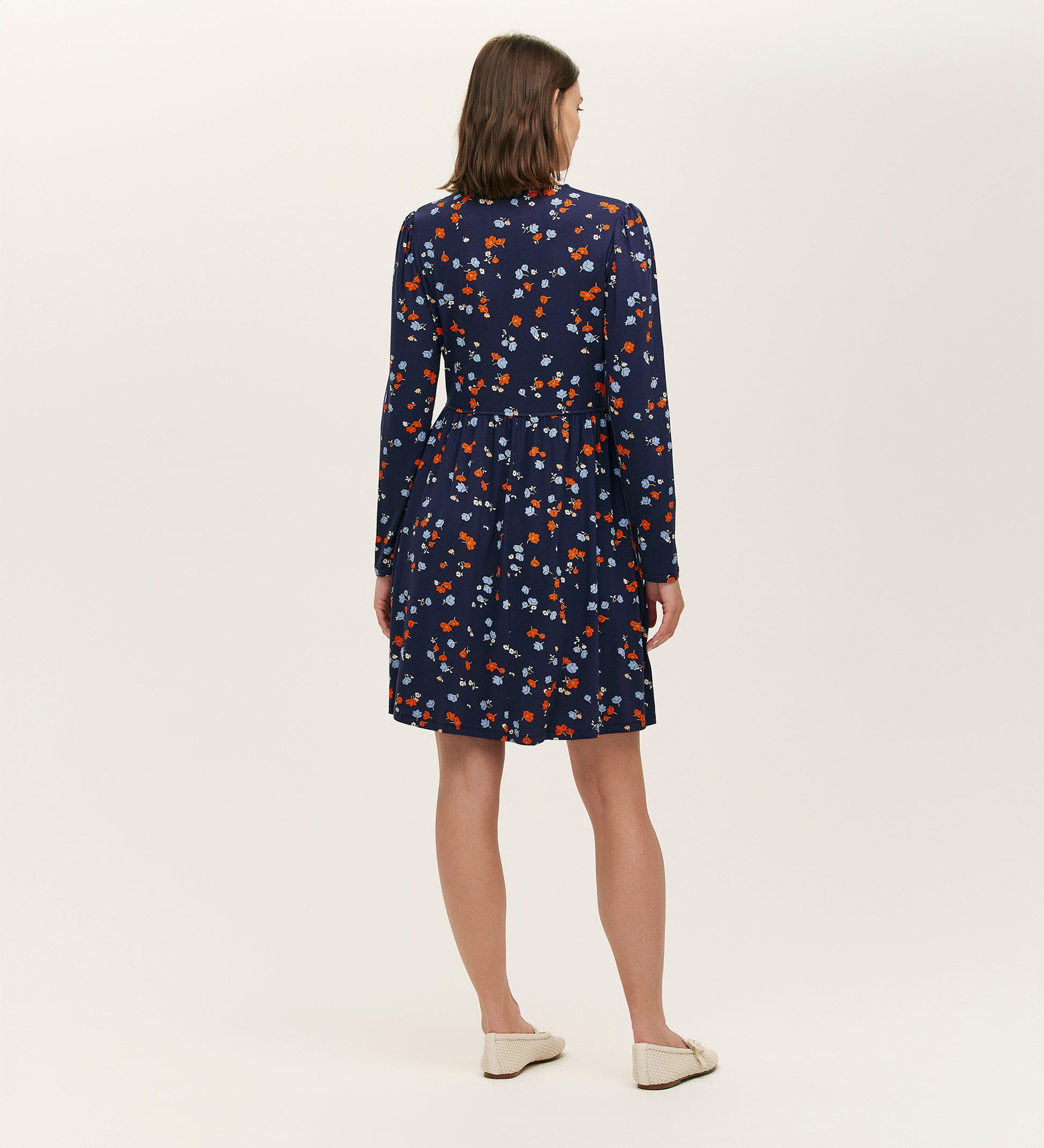 A woman with shoulder-length brown hair stands facing away, wearing the Joanne Navy Ditsy Jersey Short Dress featuring a V-neck and long sleeves. She pairs it with white flats against a plain, light-colored background.