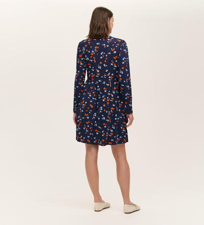 A woman with shoulder-length brown hair stands facing away, wearing the Joanne Navy Ditsy Jersey Short Dress featuring a V-neck and long sleeves. She pairs it with white flats against a plain, light-colored background.