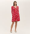 A woman stands against a plain background wearing the Joanne Red Poppies Jersey Short Dress, featuring long sleeves and a white floral pattern. She has her hands in the pockets and pairs it with white flat shoes for everyday comfort.