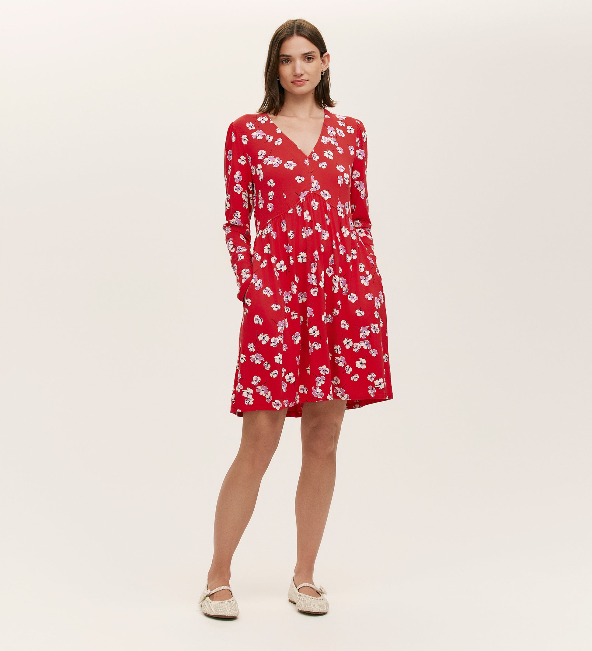 A woman stands against a plain background wearing the Joanne Red Poppies Jersey Short Dress, featuring long sleeves and a white floral pattern. She has her hands in the pockets and pairs it with white flat shoes for everyday comfort.