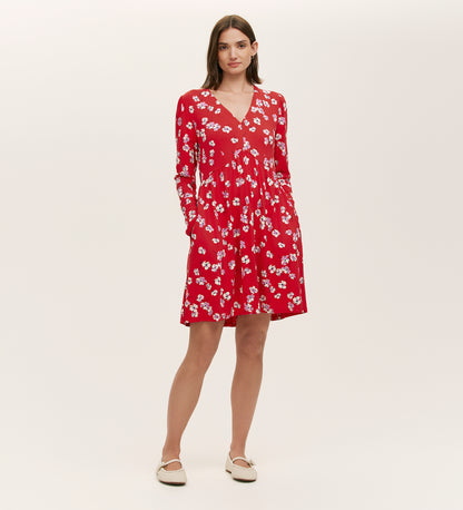 A woman stands against a plain background wearing the Joanne Red Poppies Jersey Short Dress, featuring long sleeves and a white floral pattern. She has her hands in the pockets and pairs it with white flat shoes for everyday comfort.