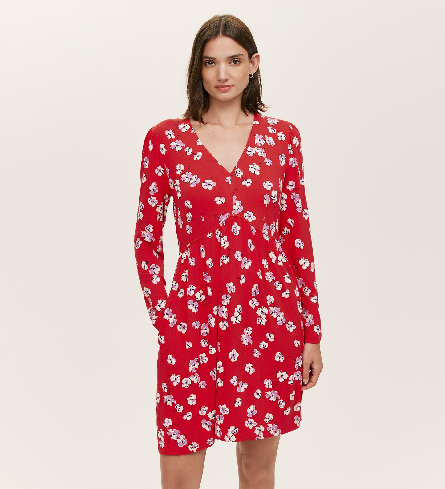 A woman in the Joanne Red Poppies Jersey Short Dress, featuring long sleeves, a V-neckline, and a white floral print, stands against a plain light background, embodying effortless everyday comfort.
